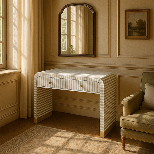 A pale gray bone inlay 2-drawer waterfall style desk with brass handles, placed in a room with a window and a chair.