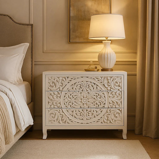 A white, hand-carved wooden Lombok 3-drawer dresser with intricate designs, placed next to a bed with a lamp on top.